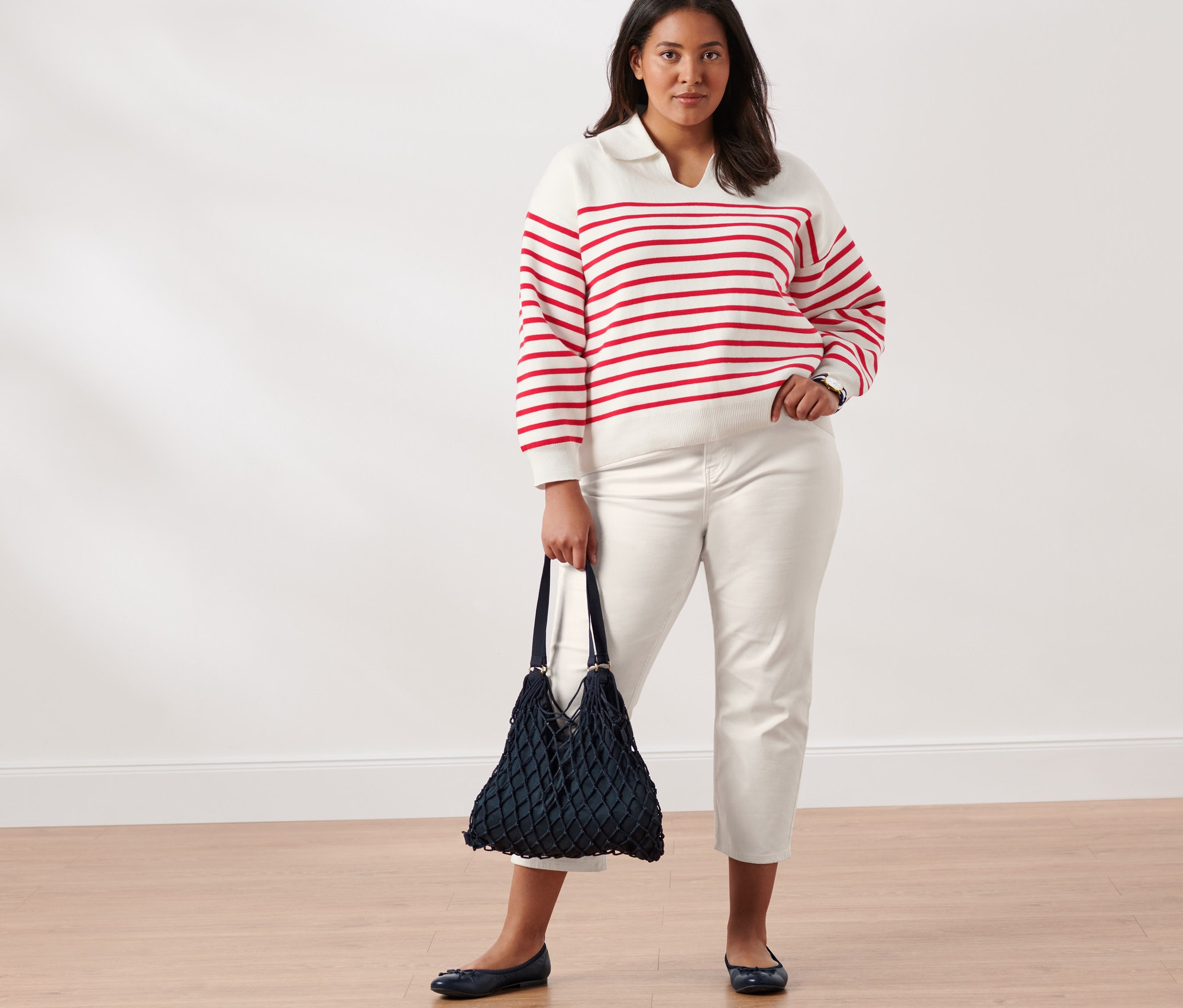 Žena pózuje v pletenom pulóvri s golierom bielo-červenej farby, džínsoch Slimfit, kožených balerínach, so sieťovou taškou a náramkovými hodinkami z ušľachtilej ocele s quartzovým strojčekom.