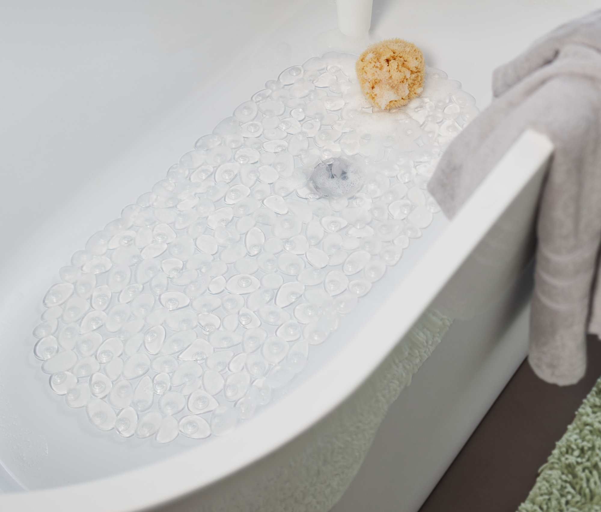 Vo vani je priehľadná podložka do vane. Na okraji vane visí sivý uterák a vedľa vane leží zelený koberec.