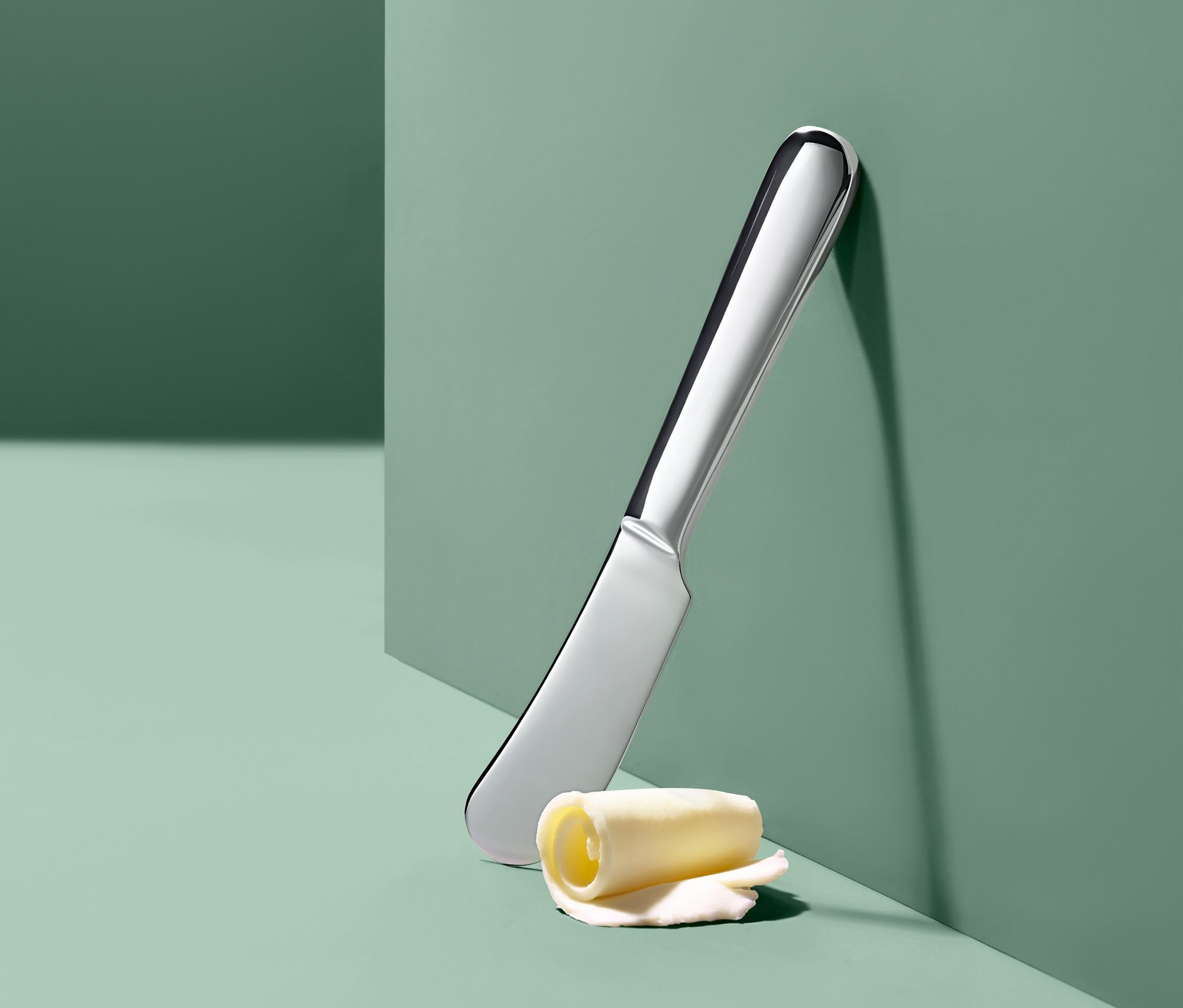 Strieborný nôž na maslo a rolka masla opreté o zelenú stenu.