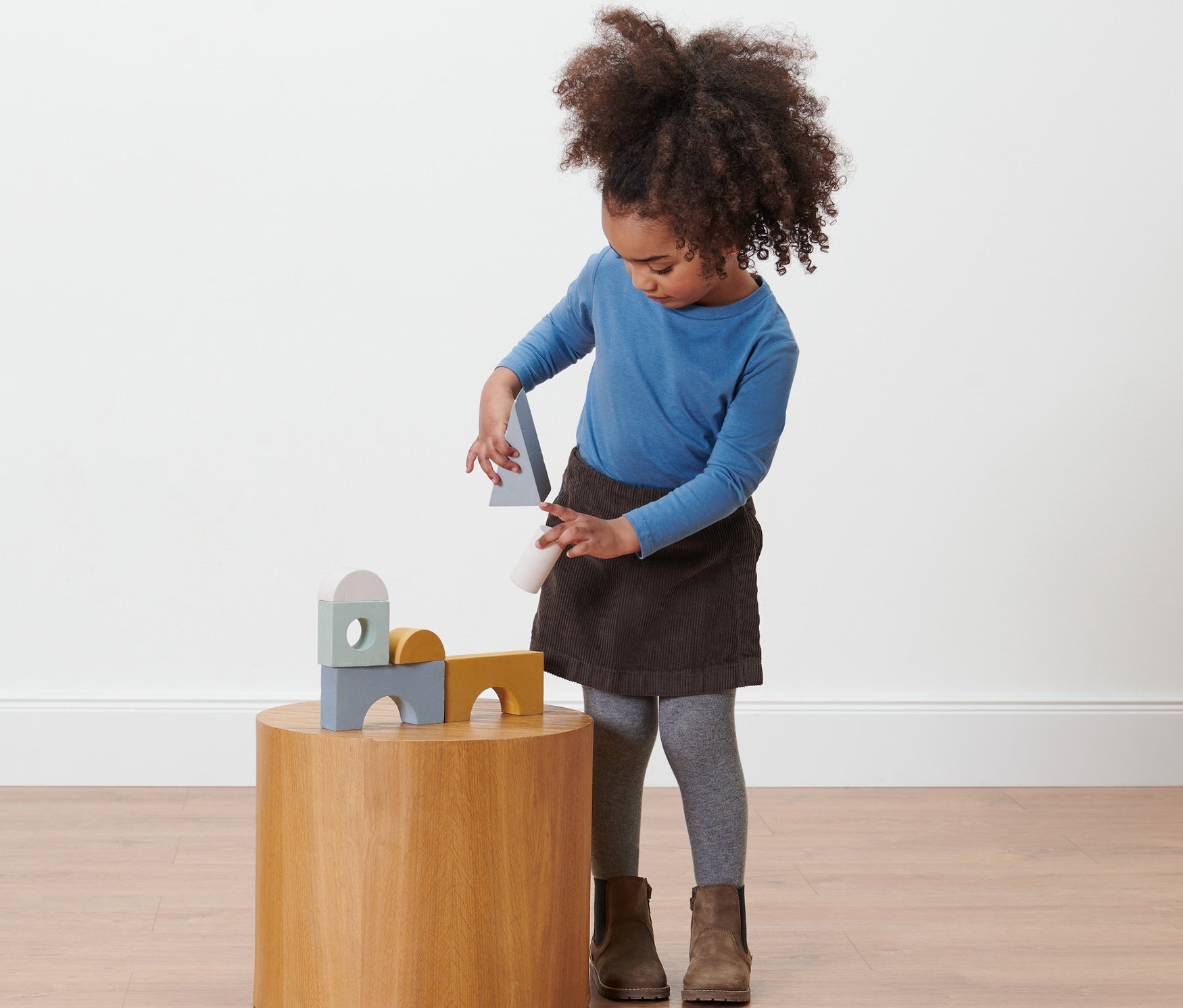 Dievča sa hrá s drevenými hračkami na okrúhlom stolíku, má na sebe detskú nohavicovú sukňu.