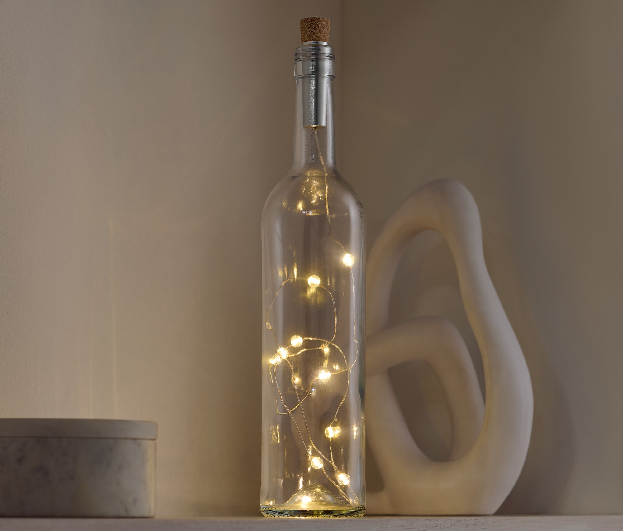 Dekoratívna sklenená fľaša s korkovou zátkou, vnútri ktorej sú svetlá, stojí na poličke vedľa ozdobnej sošky a šedej krabičky.
