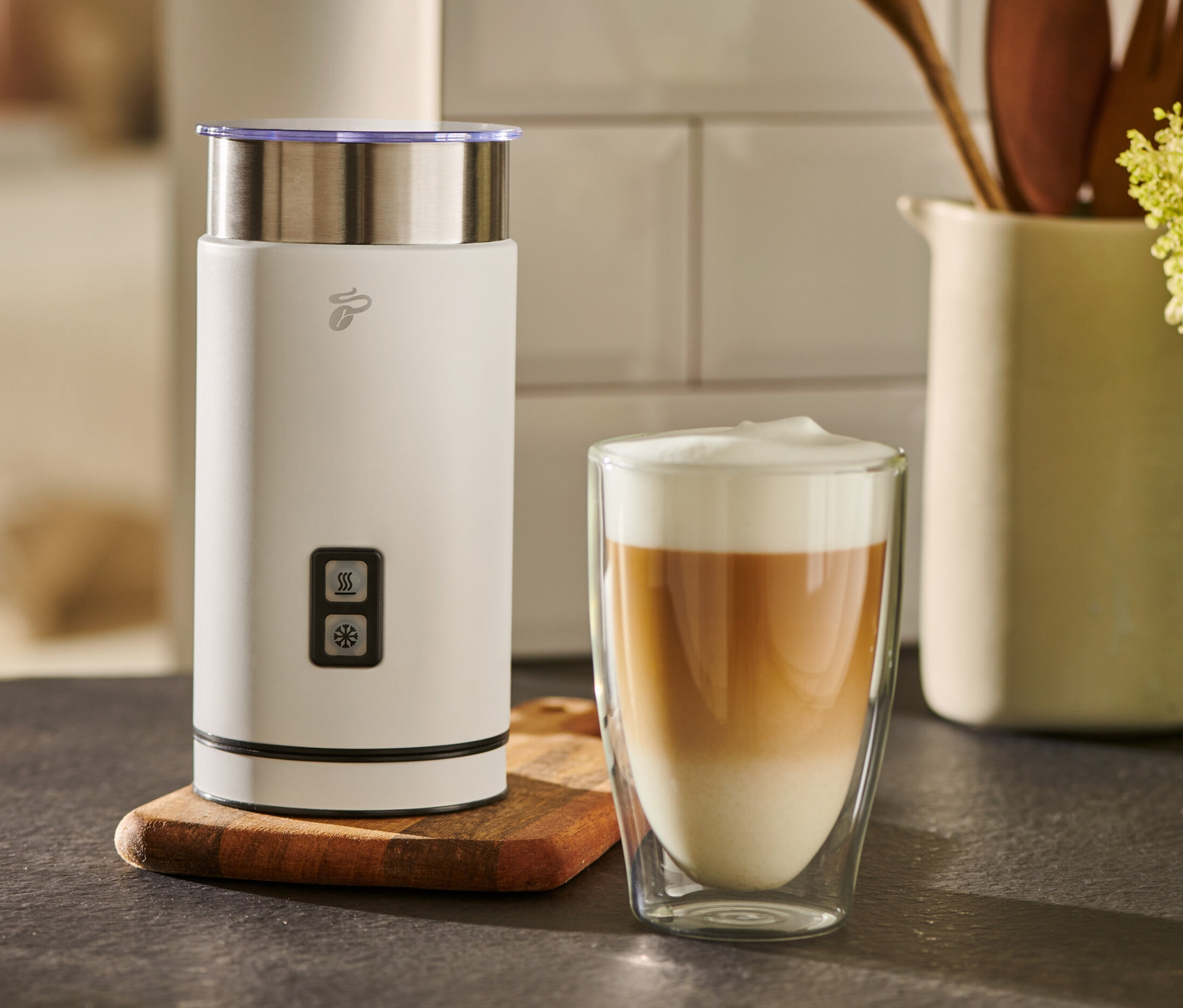 Biely elektrický napeňovač mlieka stojí vedľa pohára s kávou s mliekom.