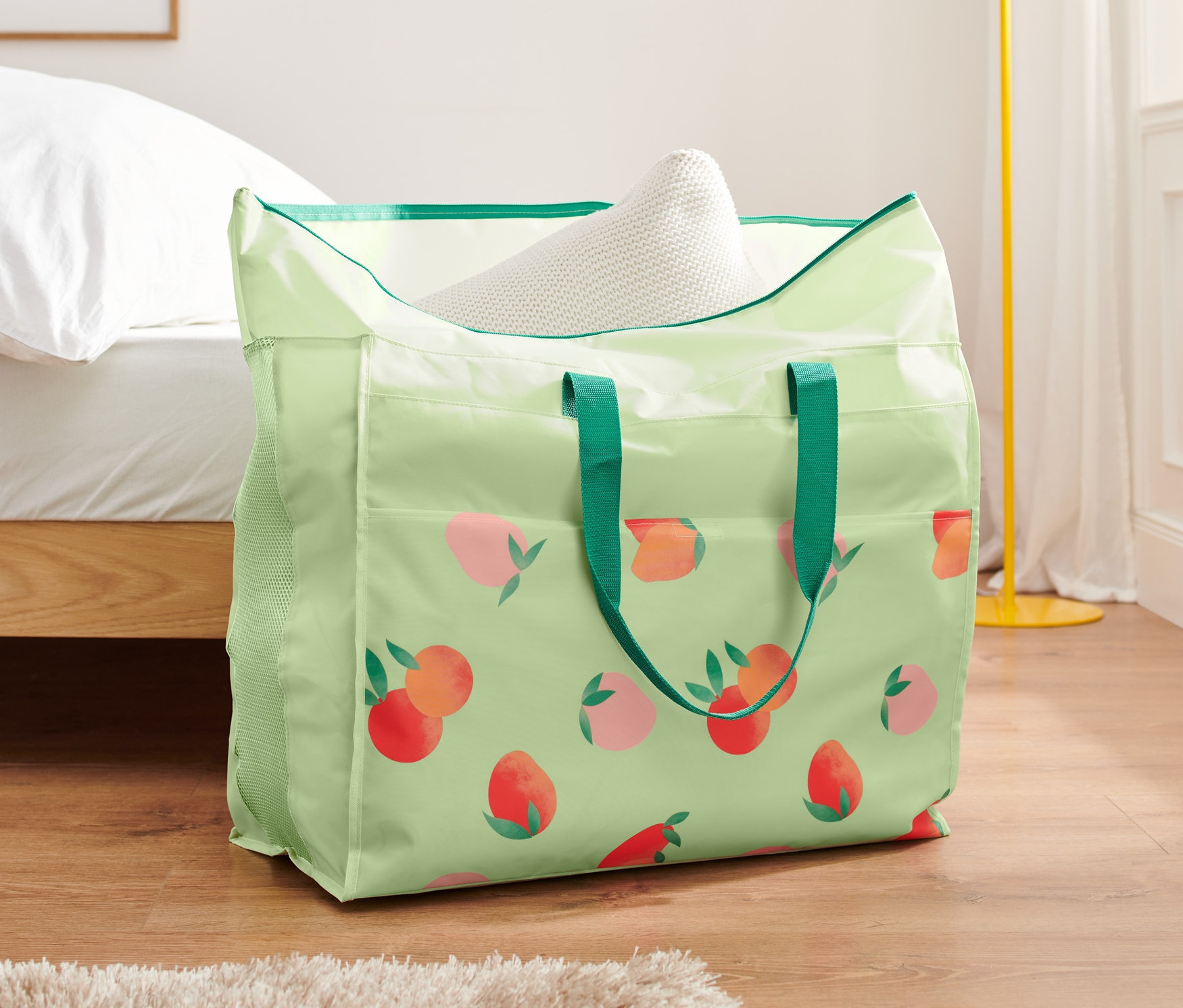 Zelená taška na lôžkoviny s potlačou broskýň stojí na podlahe pri posteli.