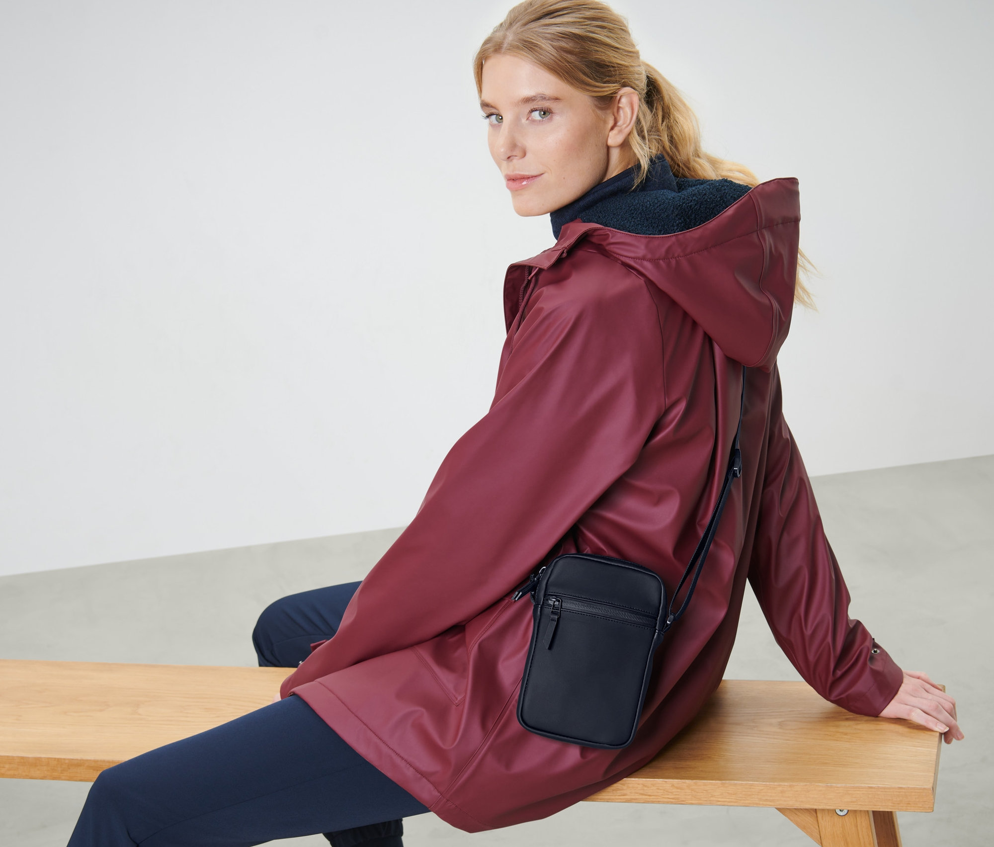 Blondínka sedí na lavici v bordovej bunde a s čiernou taškou na plece do dažďa.