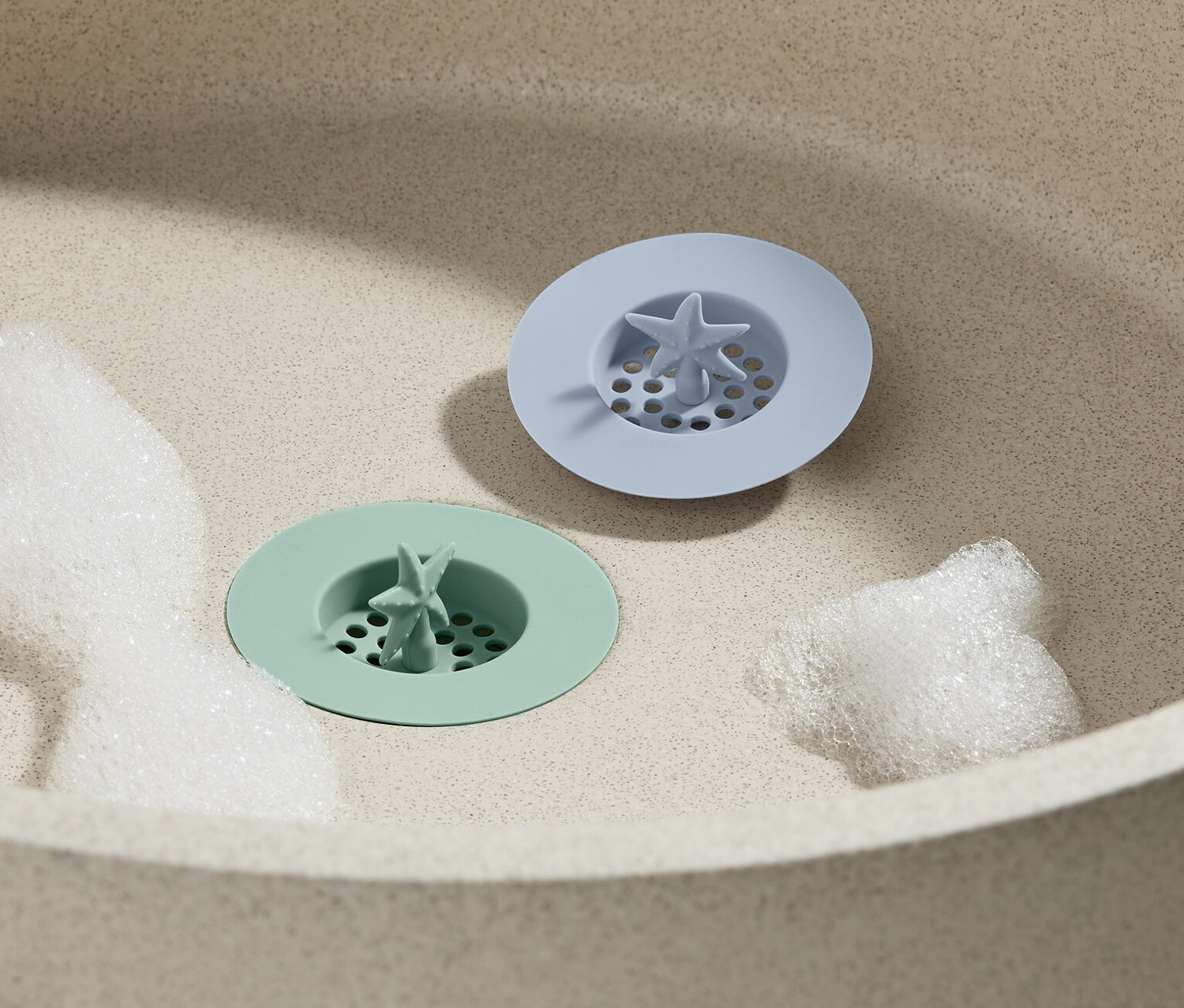 V umývadle ležia dve silikónové sitká do odtoku, 2 ks, jedno zelené a jedno modré, obklopené penou.