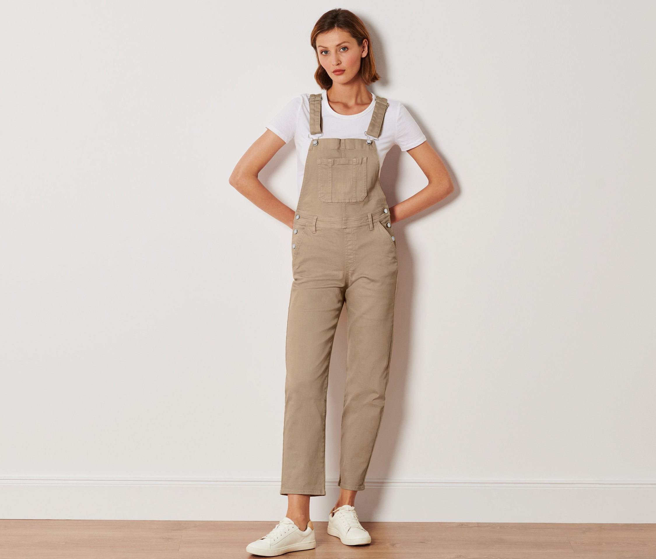 Žena pózuje v béžových nohaviciach na traky, bielom tričku a bielych teniskách.