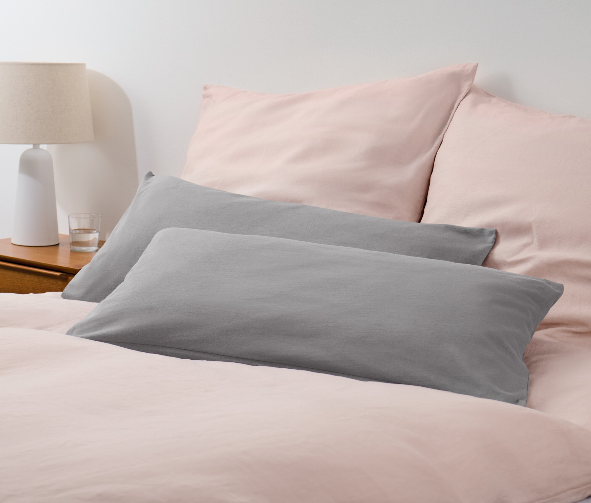 Dve sivé džersejové obliečky na vankúše ležia na posteli s ružovou posteľnou bielizňou.