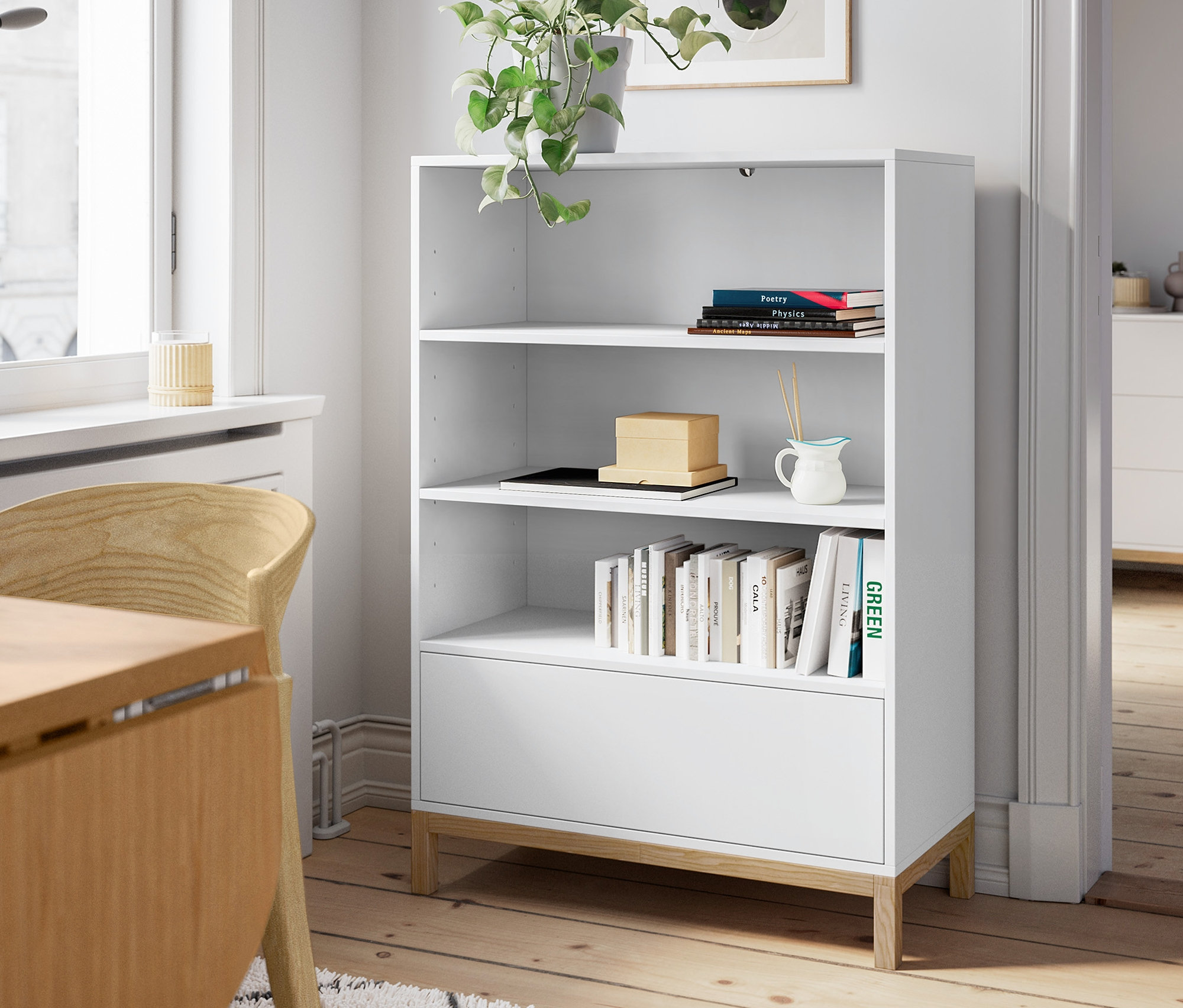 Biely regál stojí vedľa okna a dreveného jedálenského stola. Na regáli je rastlina a komoda je viditeľná v pozadí.