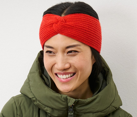 Žena so skríženými rukami sa usmieva a má na sebe oranžovú pletenú čelenku a zelenú páperovú bundu.