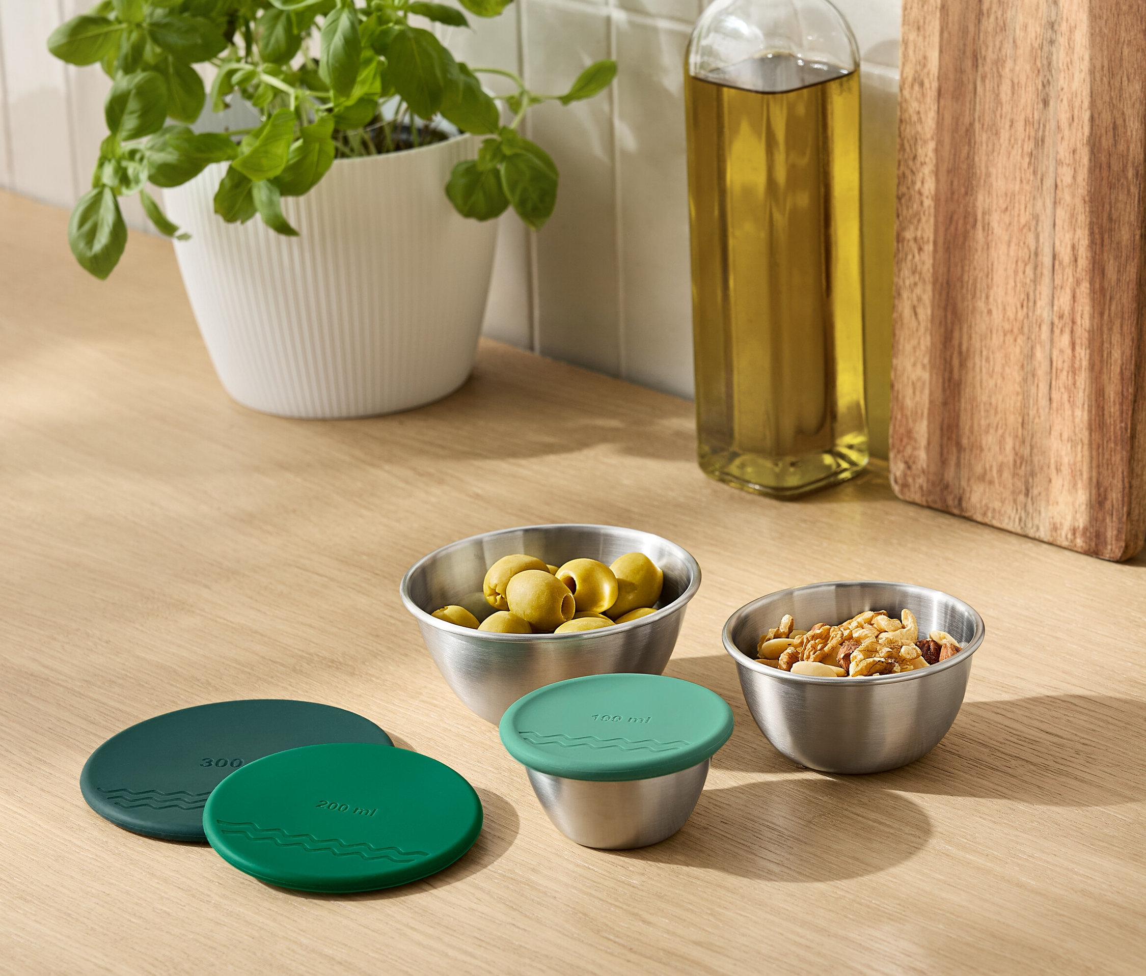 Na linke stojí rastlina v kvetináči, fľaša oleja, doska na krájanie a kovové misky s olivami a orechmi. Miska je zakrytá zeleným vekom.