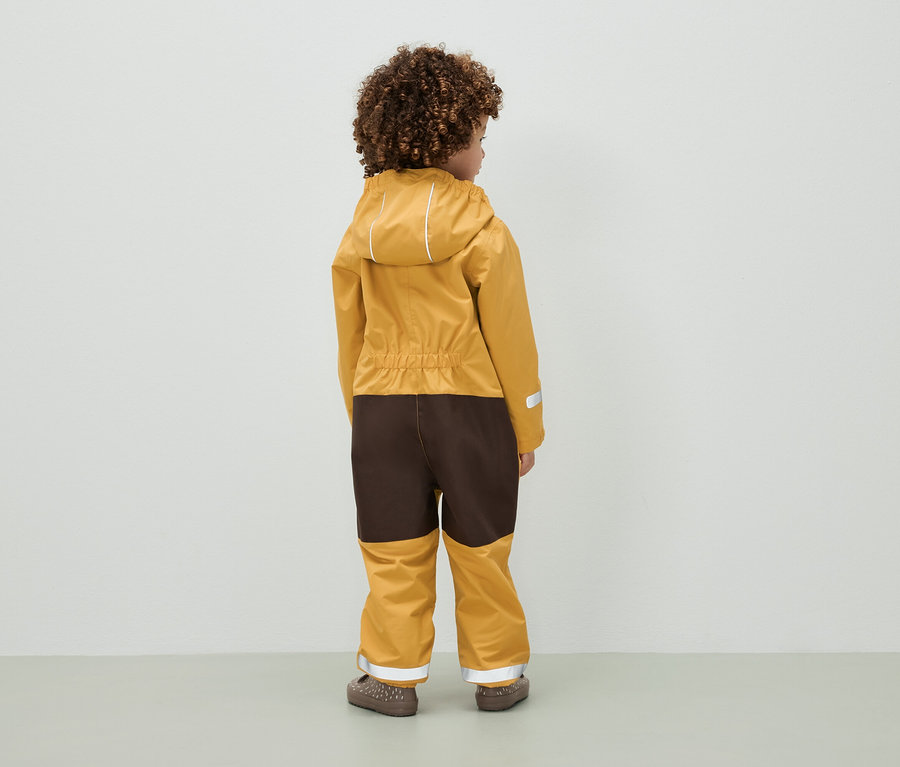 Dieťa s kučeravými vlasmi stojí chrbtom a má na sebe hnedo-horčicovú kombinézu s kapucňou.