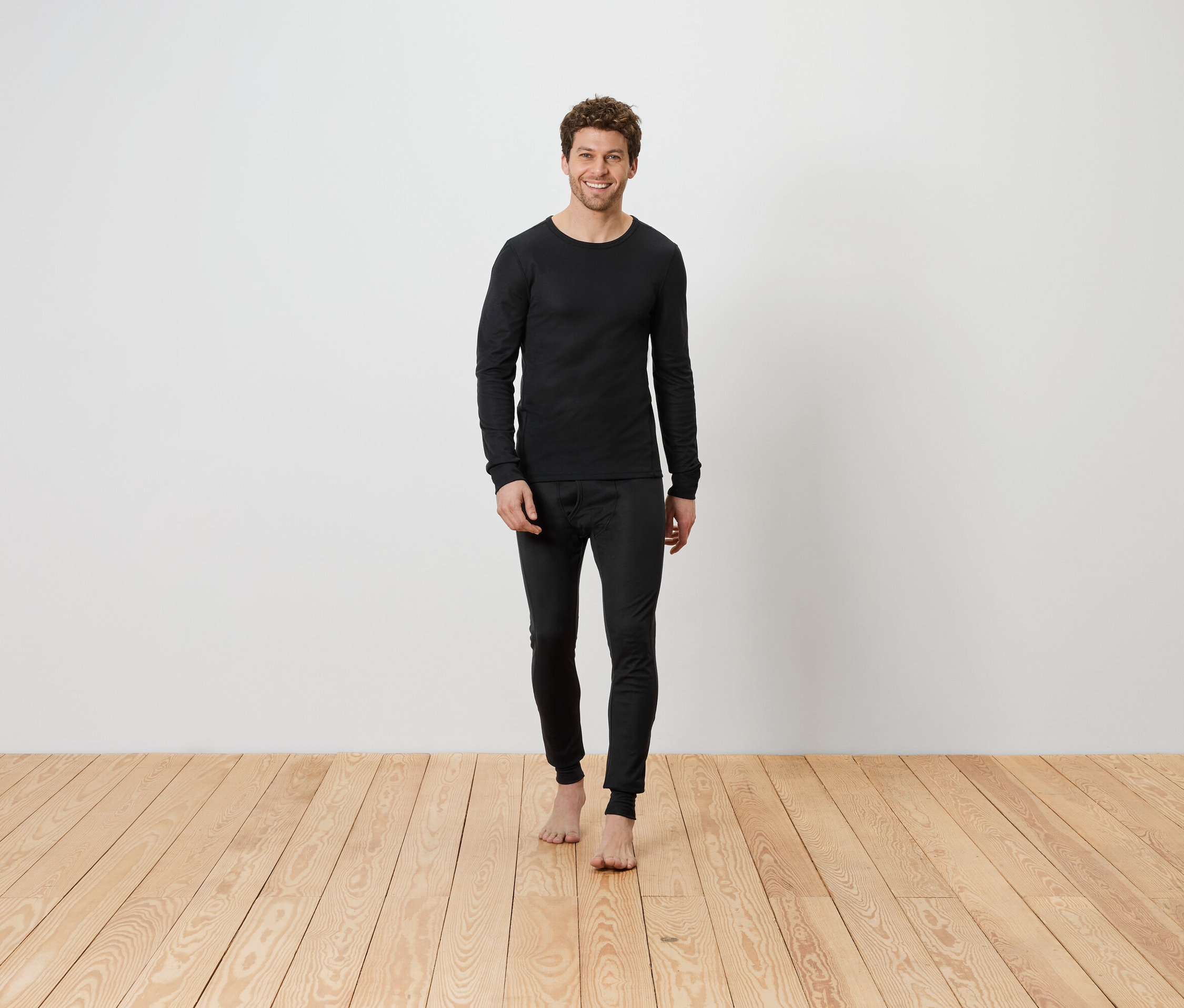 Muž v čiernej termo bielizni stojí bosý na drevenej podlahe pred bielou stenou.