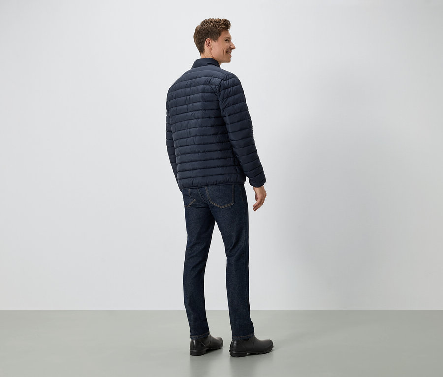 Muž stojí chrbtom k fotoaparátu a má na sebe tmavomodrú páperovú bundu, džínsy a čierne topánky.