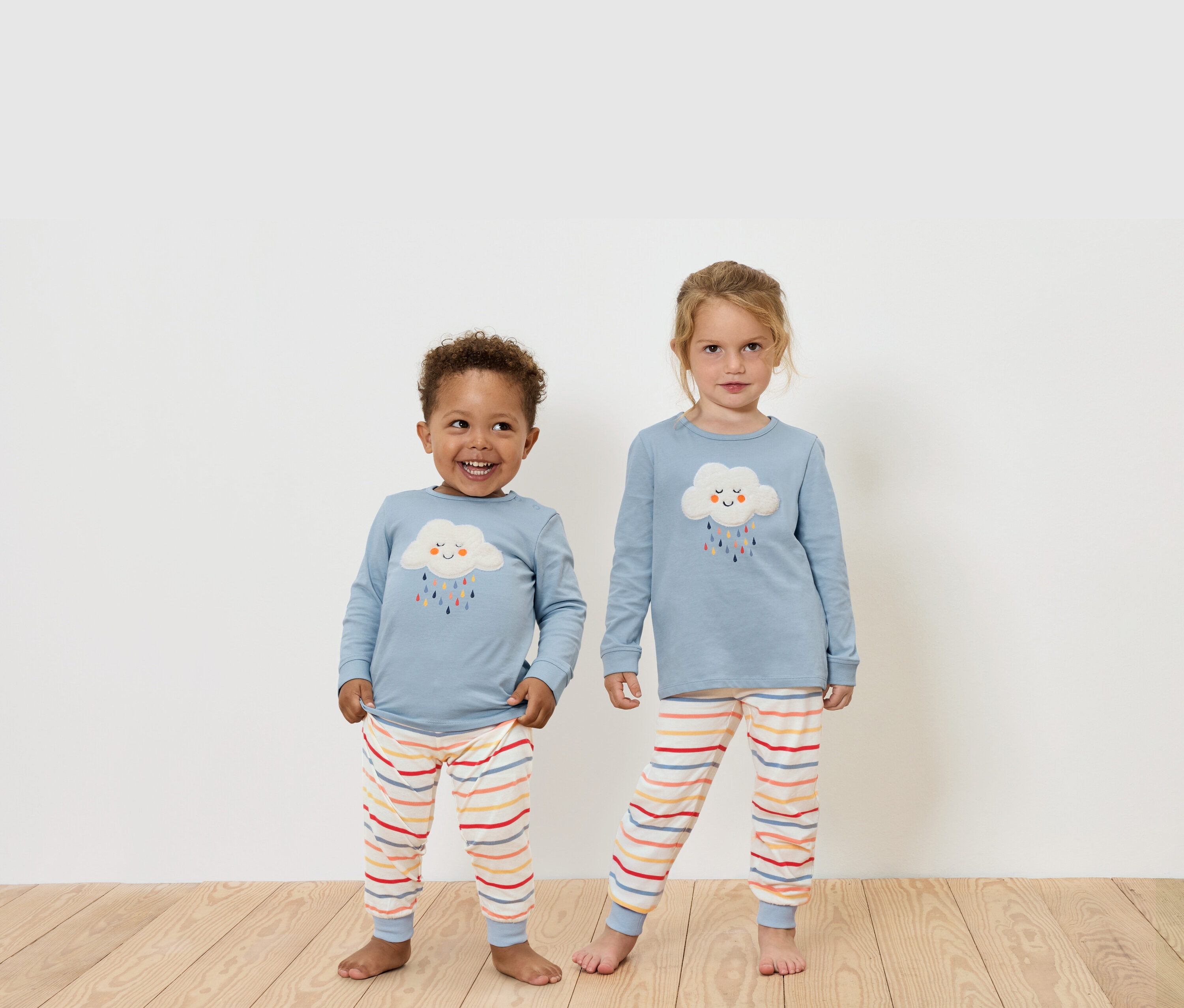 Dve deti stoja na drevenej podlahe a majú na sebe modré tričká s dlhým rukávom a pruhované nohavice.