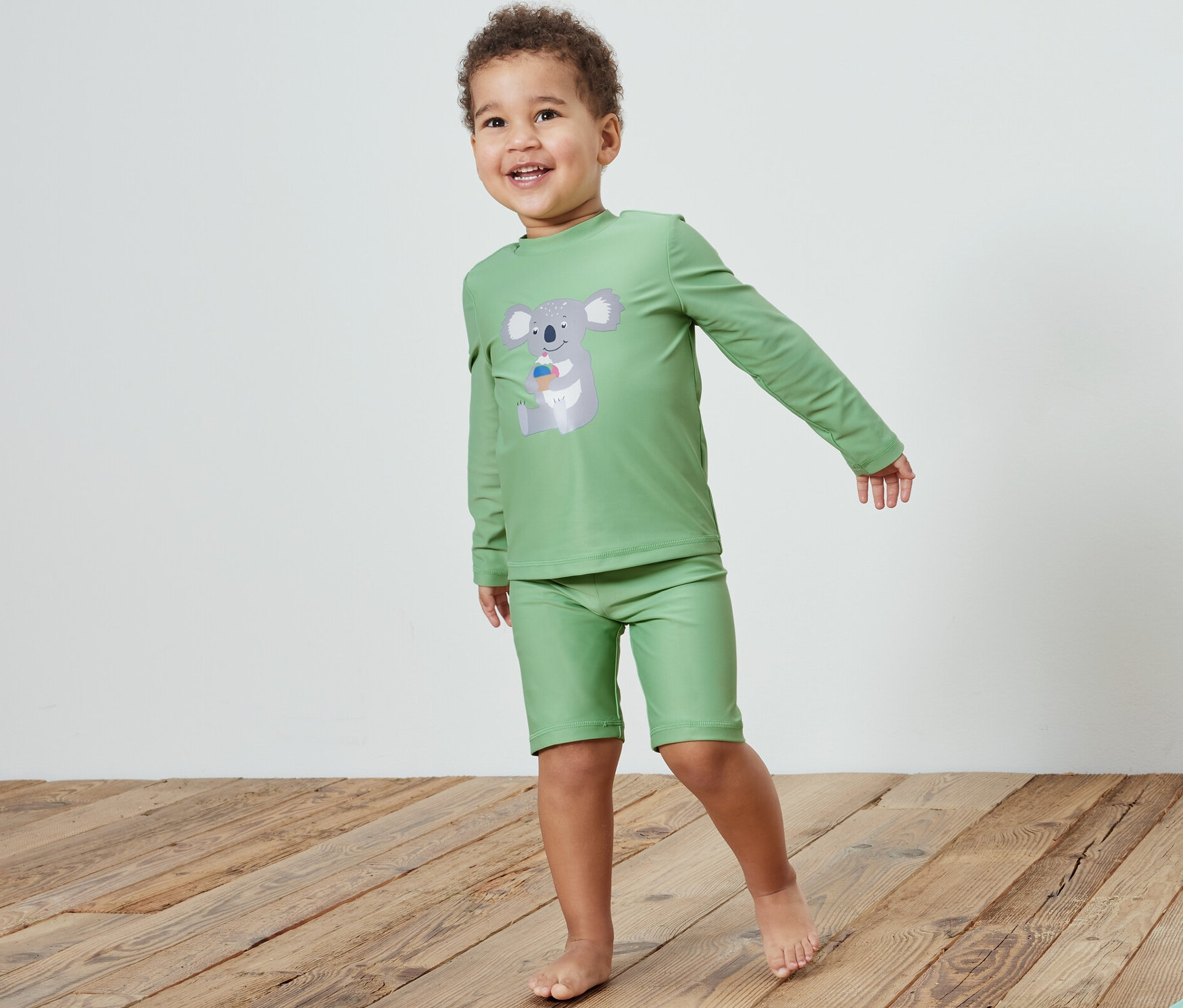 Chlapec v detskej súprave s ochranným UV faktorom 80, koala, stojí na drevenej podlahe vedľa hračky.