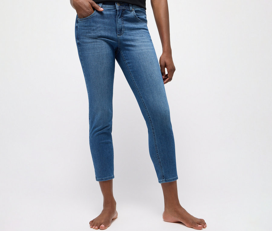 Osoba má na sebe modré džínsy Angels »Ornella« strih Slim Fit 7/8 s jednou rukou vo vrecku, bosá.
