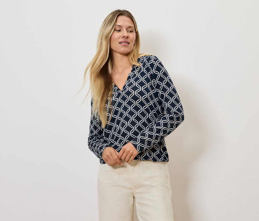 Blondínka pózuje v blúzke s geometrickým vzorom a svetlých nohaviciach.
