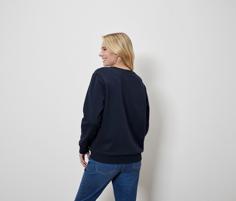 Blondínka sa pozerá cez rameno, má na sebe modrú mikinu a džínsy.