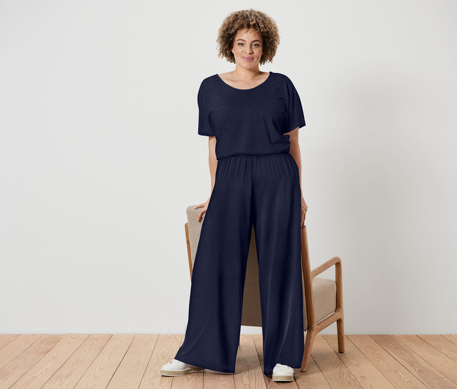 Žena s kučeravými vlasmi má na sebe tmavomodrý overal a biele topánky, stojí vedľa stoličky s dreveným rámom.