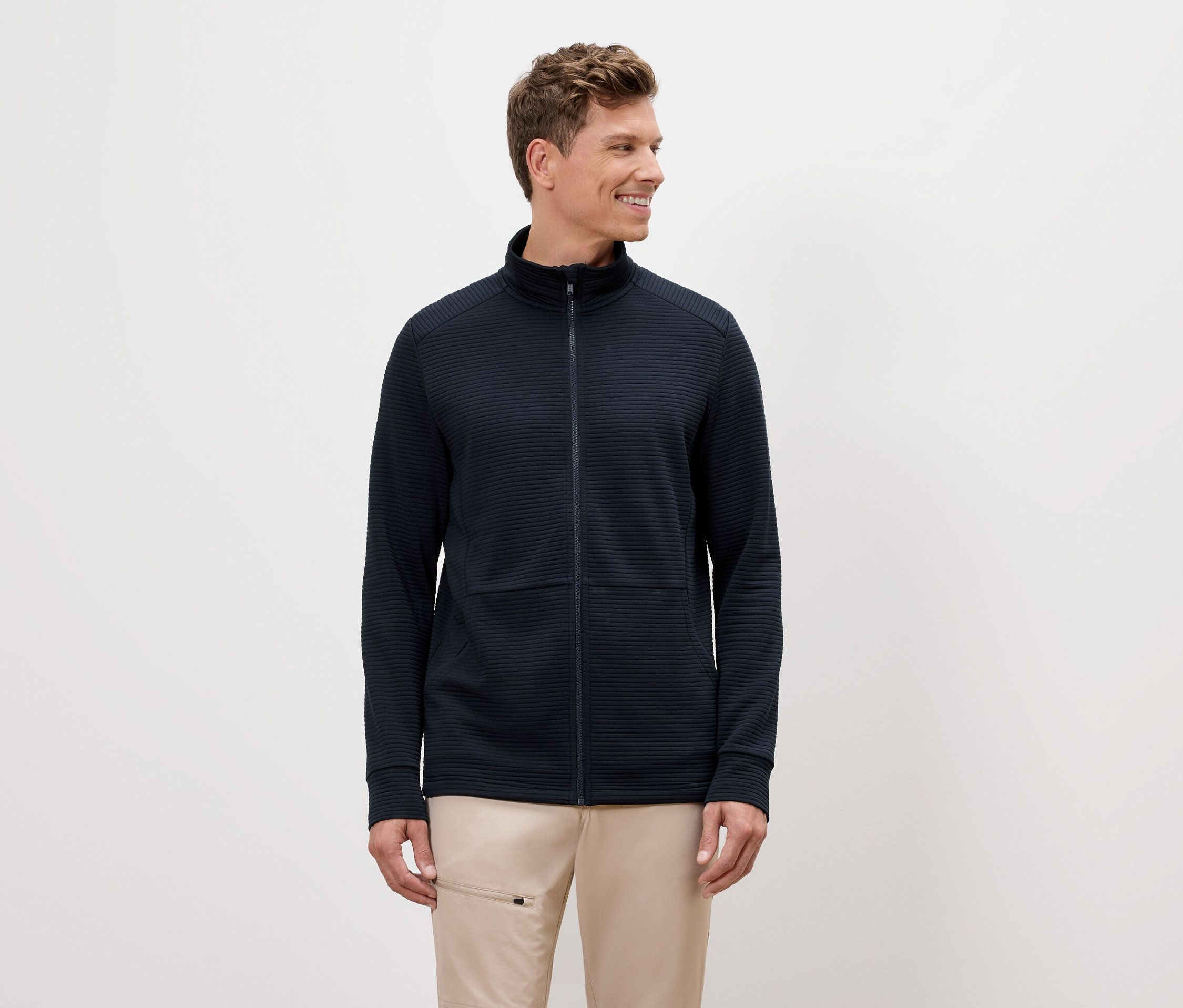 Muž s blond vlasmi má na sebe tmavomodrú bundu na zips a béžové nohavice.