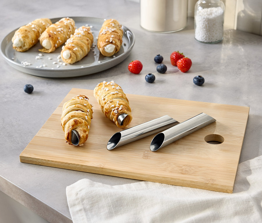 Na stole sú trubičky s krémom na tanieri a doske na krájanie, spolu s bobuľami, cukrom a kovovými formičkami.