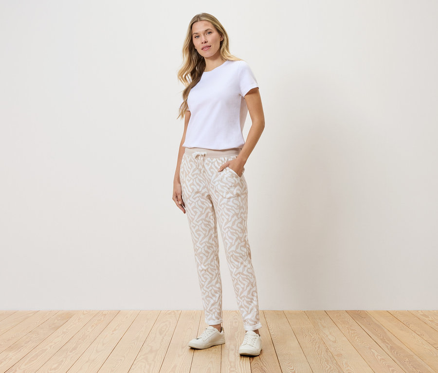Žena stojí pred stenou, má na sebe biele tričko, vzorované nohavice a biele tenisky.