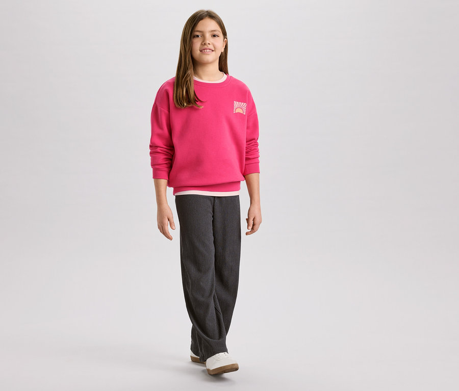 Dievča v ružovej mikine s logom východu slnka a sivých menčestrových nohaviciach, k tomu biele topánky.