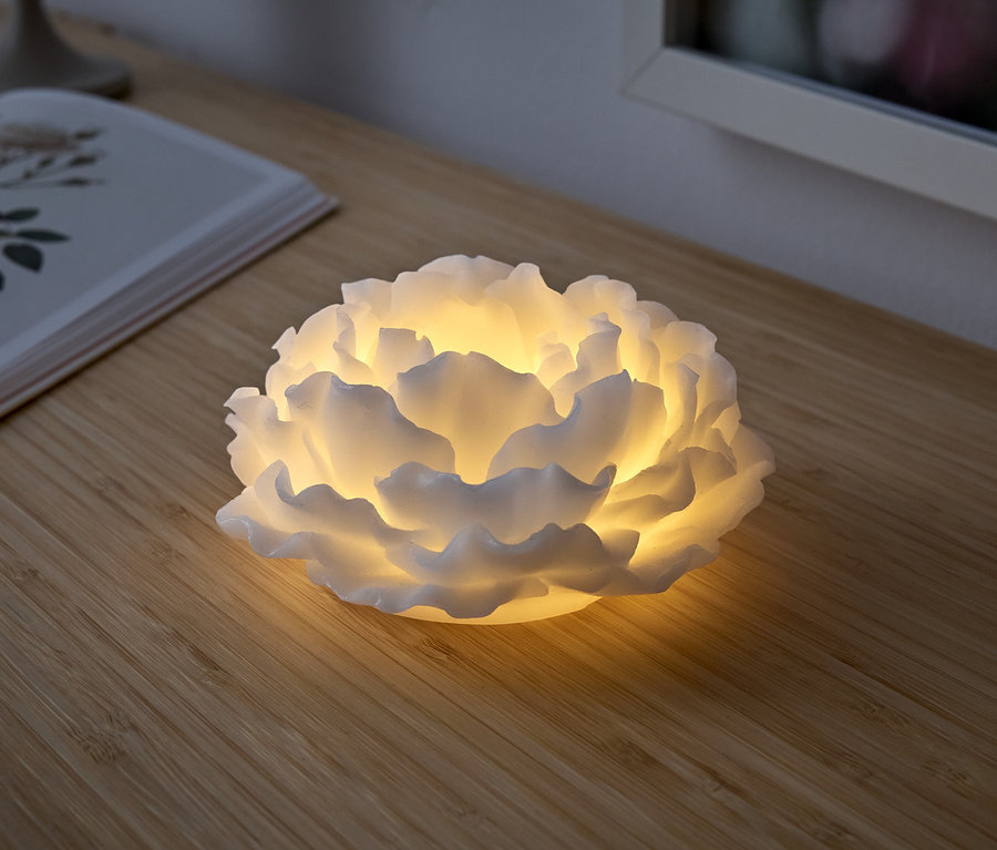 Biela lampa v tvare kvetu svieti na drevenom stole. Vedľa nej leží otvorená kniha.