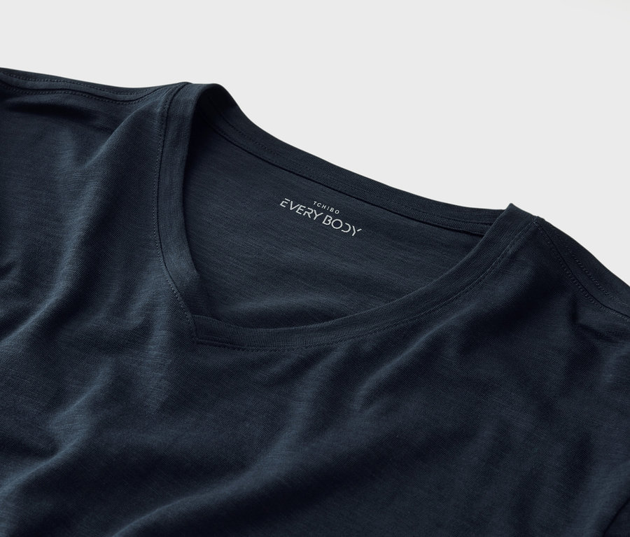 Detailný záber na tmavomodré tričko s véčkovým výstrihom a nápisom „Tchibo EVERY BODY“ pod golierom.