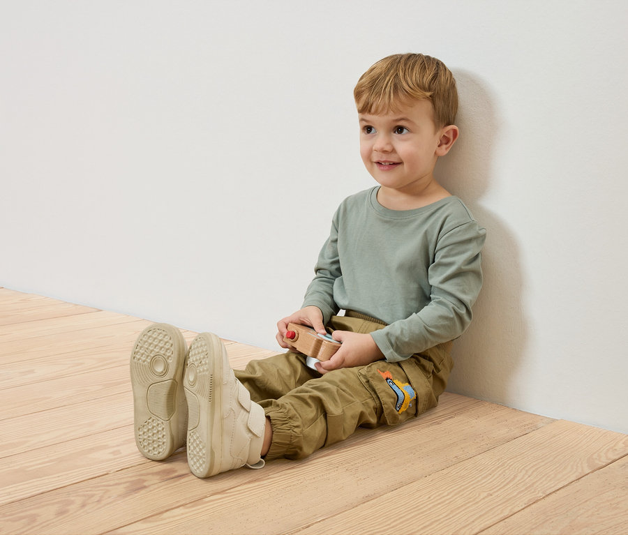 Usmievavý chlapec sedí na drevenej podlahe, opretý o bielu stenu, drží drevené autíčko.