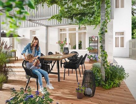 Matka a dieťa sedia pri Rozkladacom záhradnom stole s rustikálnou konštrukciou, cca 200‒250 cm. Malý a veľký Solárny lampáš s textilnou šnúrkou stojí na zemi. V pozadí je Pflanzwand mit flexibel einhängbaren Regalen »Elin«.