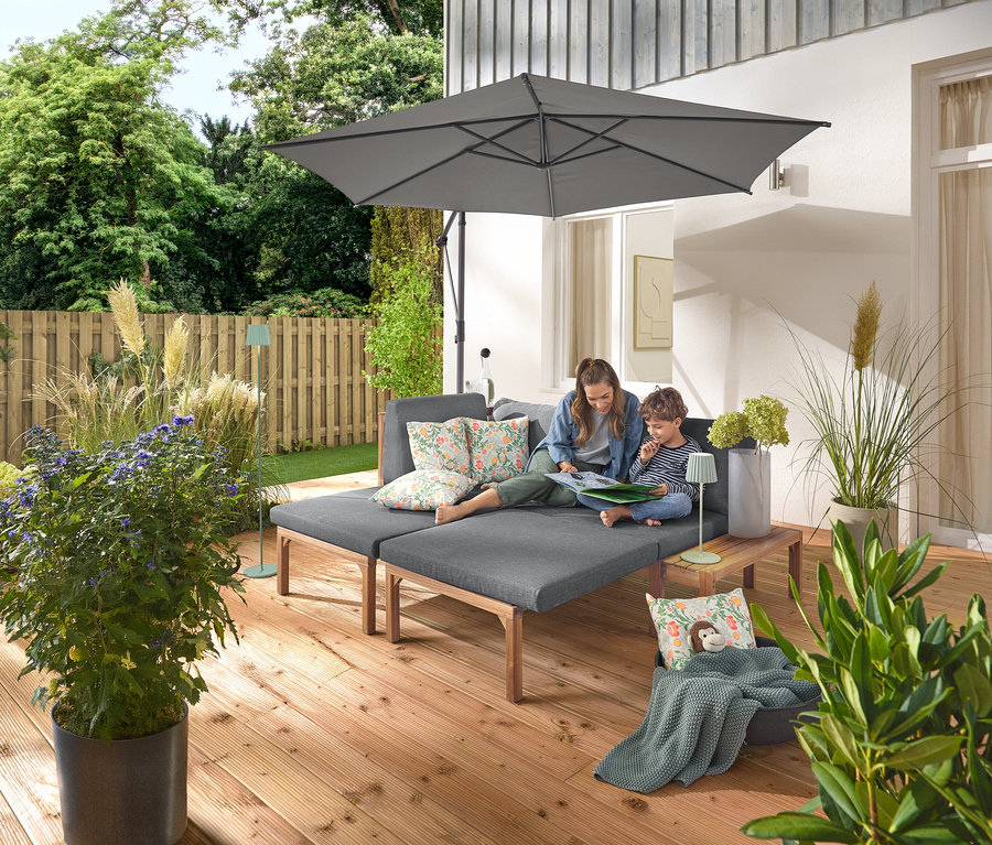 Žena a dieťa sedia na sivej záhradnej pohovke so samostatne výsuvnými podnožkami. Na pohovke sú dva vonkajšie vankúše s kvetinovým vzorom. Antracitový slnečník Leco so stojanom a krytom poskytuje tieň. Na drevenom stolíku vedľa pohovky stojí šalviovo zelená LED akumulátorová vonkajšia stolná lampa a v pozadí je vidieť šalviovo zelená LED akumulátorová vonkajšia stojacia lampa.
