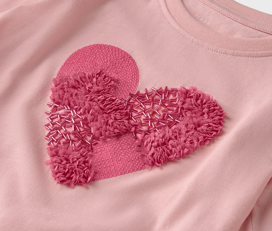 Detailný pohľad na ružové tričko s nášivkou v tvare srdca zdobenou strapcami a korálkami.