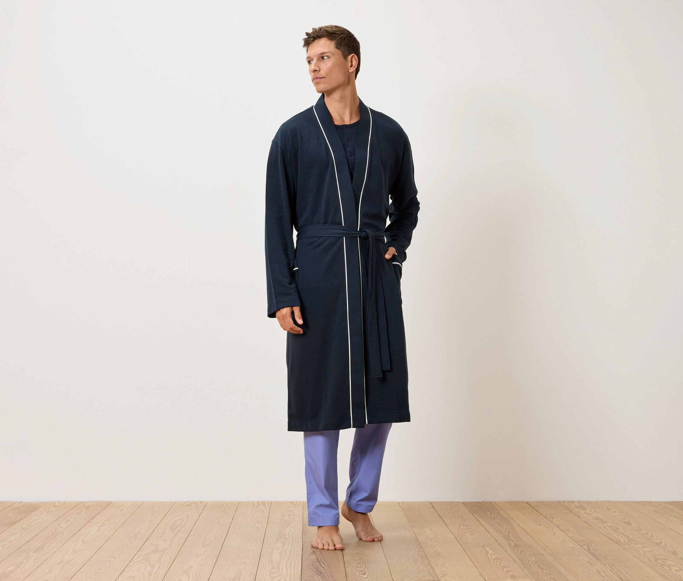 Muž stojí bosý na drevenej podlahe, oblečený v tmavomodrom župane s bielym lemovaním a modrých nohaviciach.