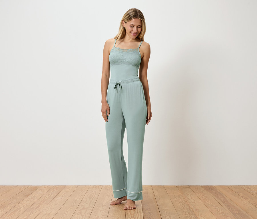 Žena má na sebe svetlozelené pyžamo s čipkovanými detailmi, stojí na drevenej podlahe pred bielou stenou.