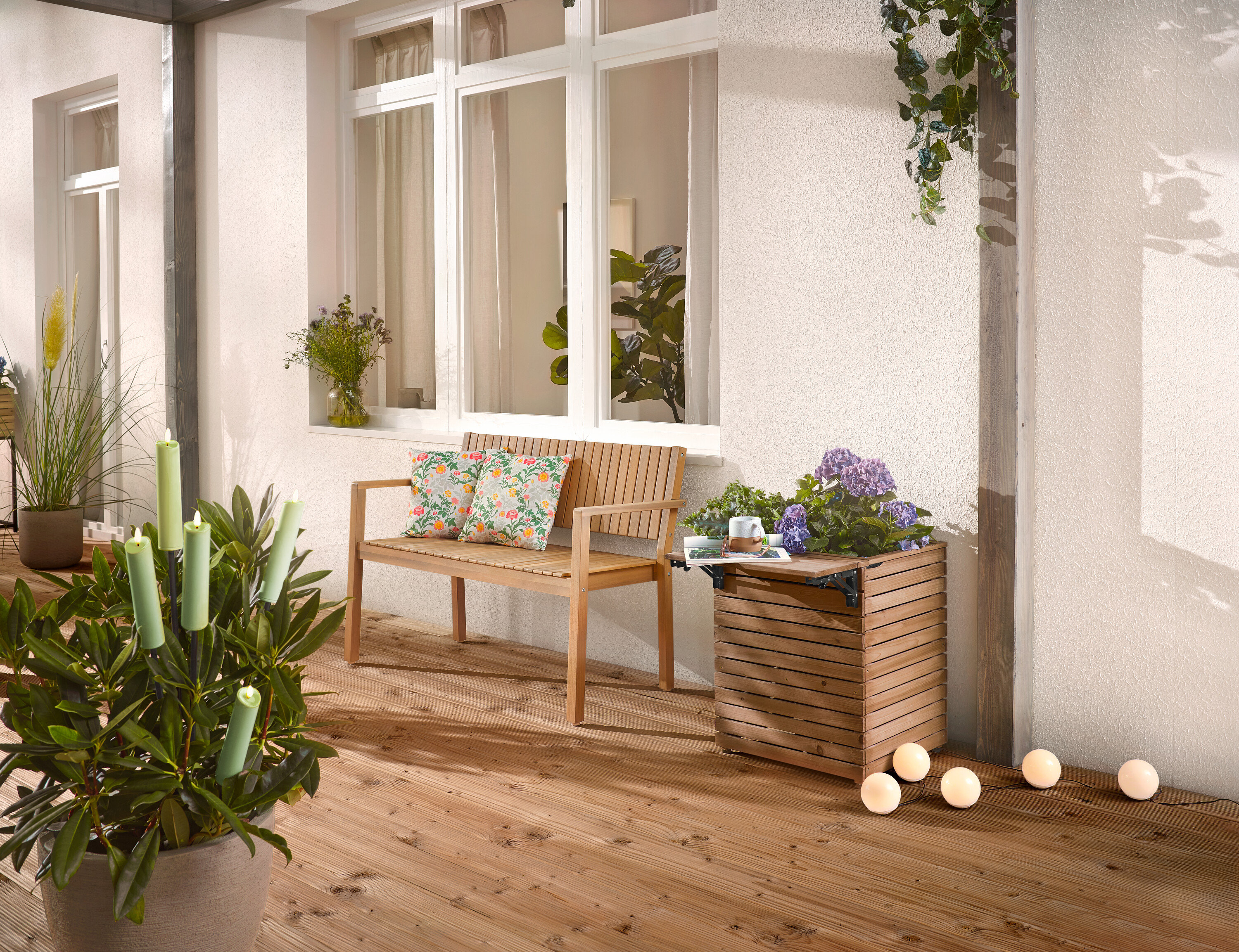 Na drevenej terase stojí drevená lavica s vankúšmi, kvetináč s kvetmi a črepníková rastlina so sviečkami.