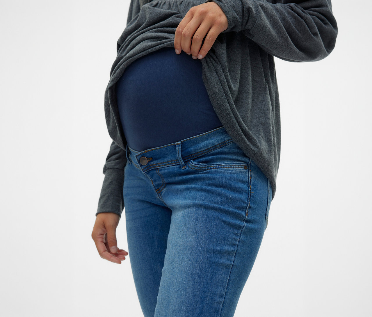 Žena si vyhrnuje sivý top nad bruchom, má na sebe skinny džínsy MAMALICIOUS »MLMILA« a tmavomodrý tehotenský top.