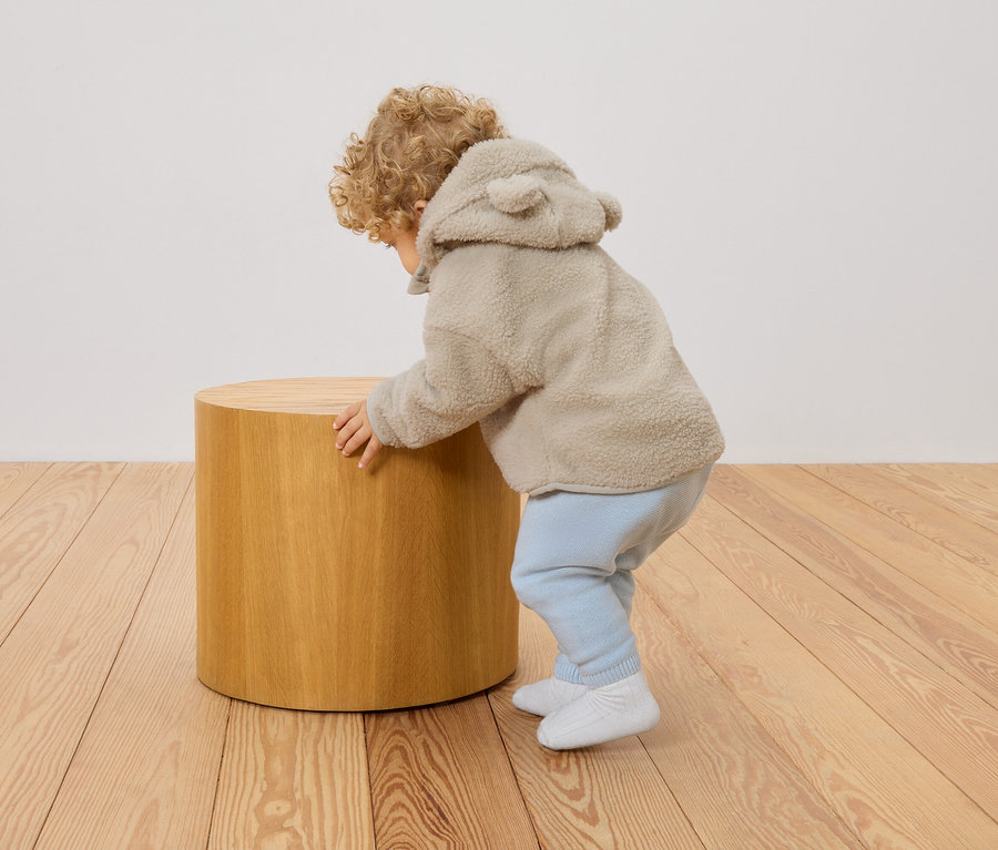 Malé dieťa s kučeravými vlasmi v béžovej teddy bunde s kapucňou a ušami sa opiera o okrúhlu drevenú stoličku.