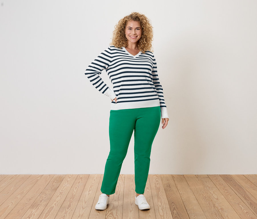 Žena s kučeravými vlasmi v modro-bielom pruhovanom svetri, zelených nohaviciach a bielych teniskách pózuje na bielom pozadí.