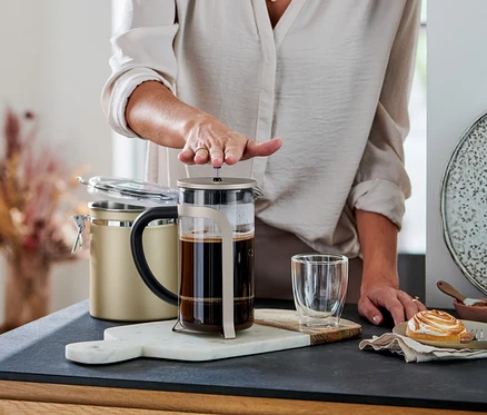 Osoba stláča piest kanvice french press (800 ml, 6 šálok, farba šampanského) naplnenej kávou. Vedľa je pohár a zákusok.