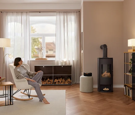 Žena sedí naboso v krémovom hojdacom kresle s látkou z buklé. Vľavo je príručný stolík s kovovými vzperami, v pozadí kovové sideboard »CN3« s palivovým drevom a vpravo policový regál s kovovými vzperami.