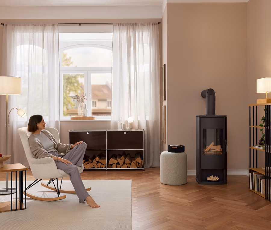 Žena sedí naboso v krémovom hojdacom kresle s látkou z buklé. Vľavo je príručný stolík s kovovými vzperami, v pozadí kovové sideboard »CN3« s palivovým drevom a vpravo policový regál s kovovými vzperami.