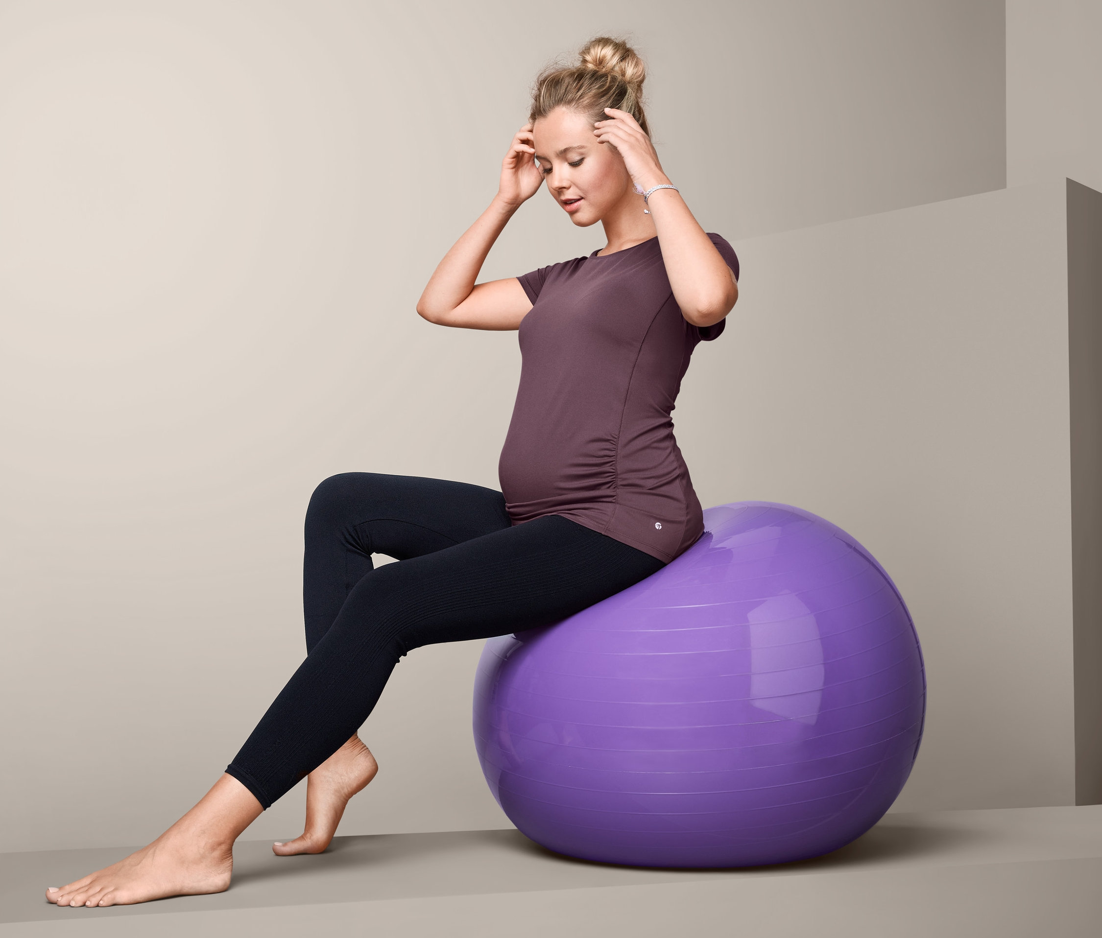 Tehotná žena sedí na fialovej gymnastickej lopte a má na sebe tehotenské športové tričko.