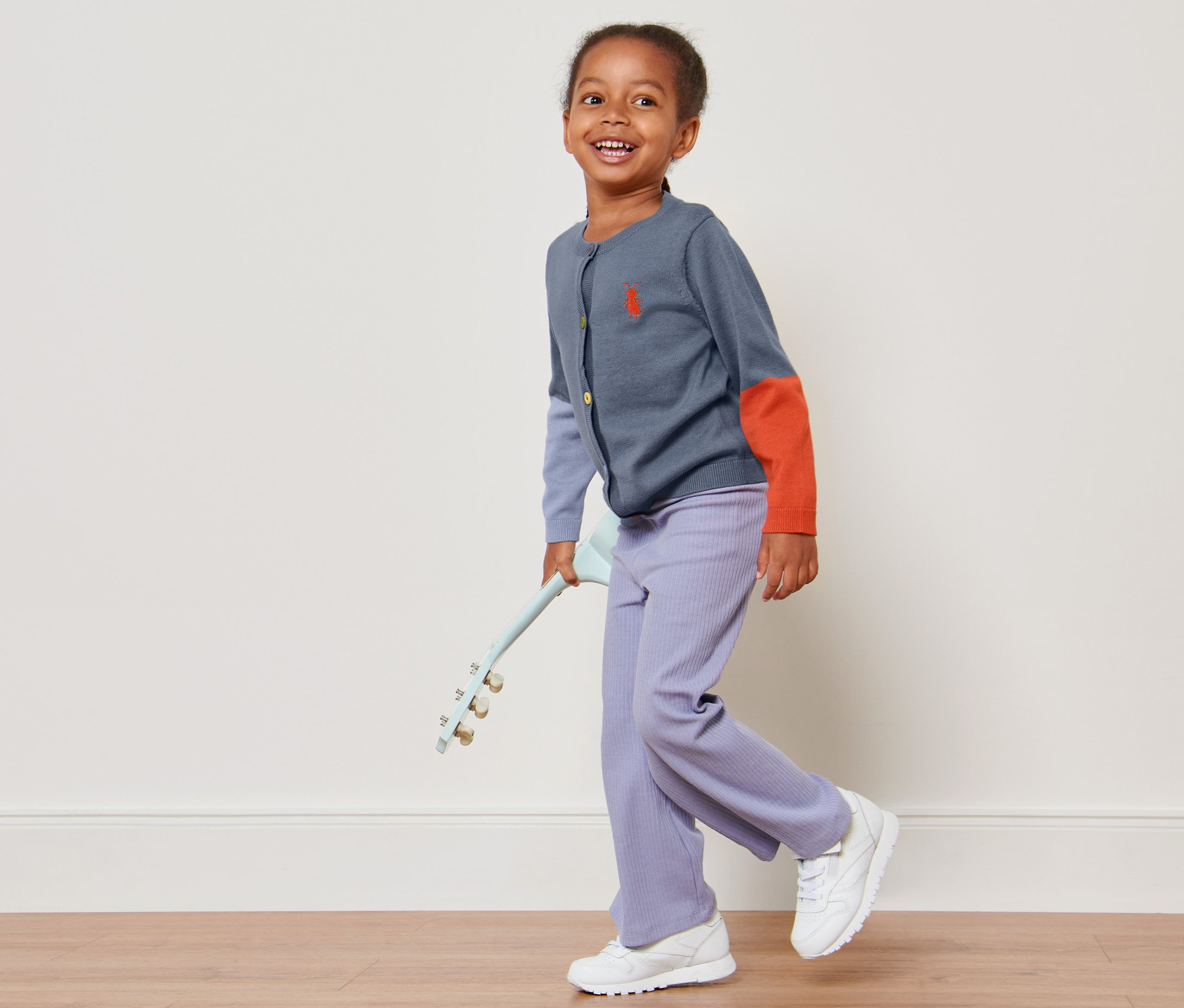 Usmievajúce sa dievčatko kráča a drží v ruke hračku gitaru, má na sebe detský pletený sveter, kombinácia kontrastných farieb.