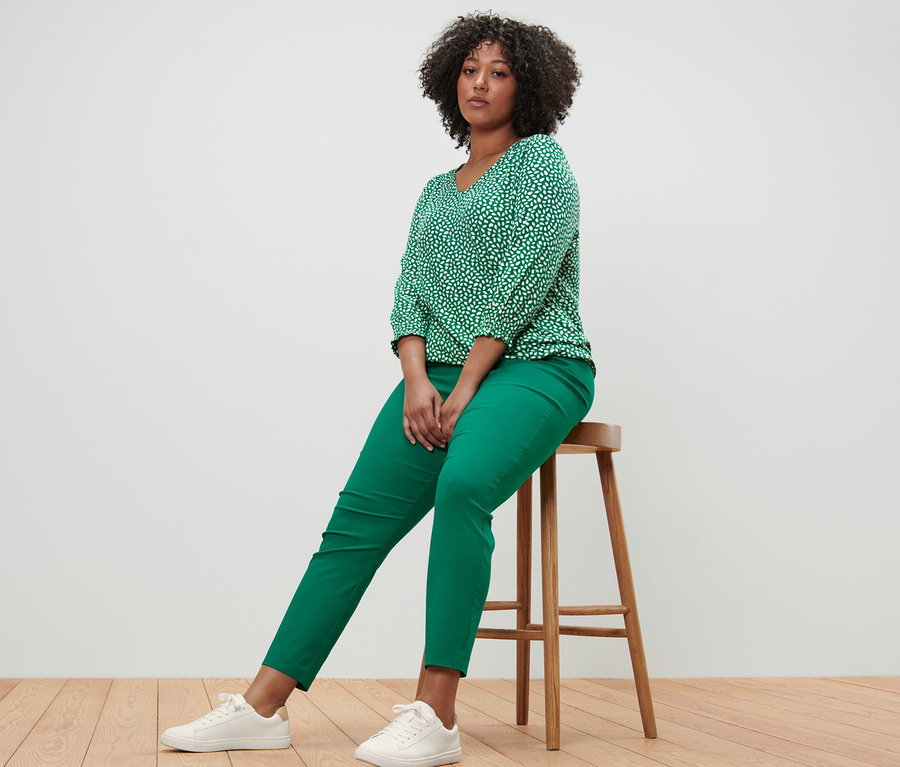 Žena sedí na stoličke a má na sebe zelené blúzkové tričko s potlačou a zelené elastické nohavice.