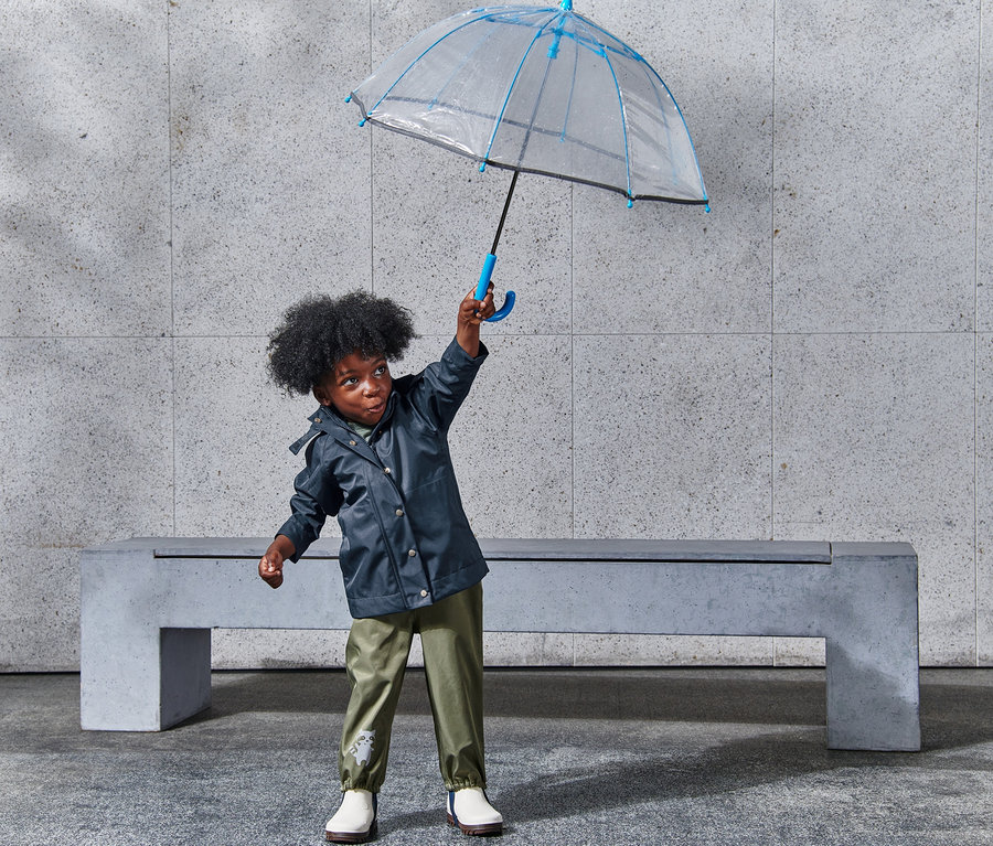 Chlapec drží nad hlavou priehľadný dáždnik s modrým lemom a má na sebe bundu do dažďa.