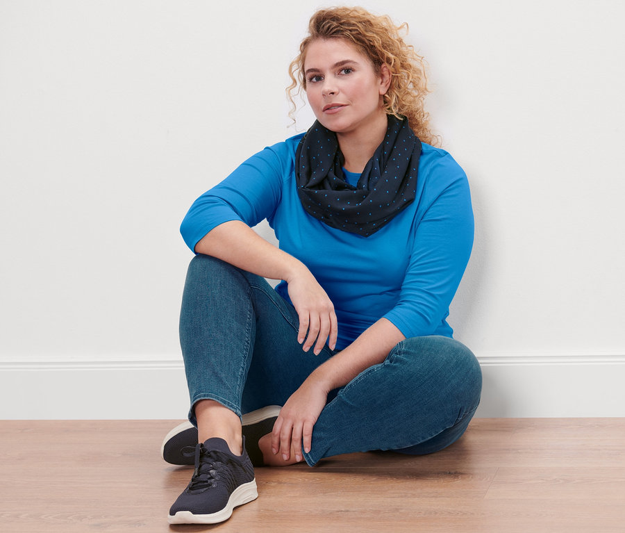 Žena sedí na podlahe a má na sebe azúrovomodré tričko s 3/4 rukávom, tregínsy Fit Nelly a bodkovaný kruhový šál.