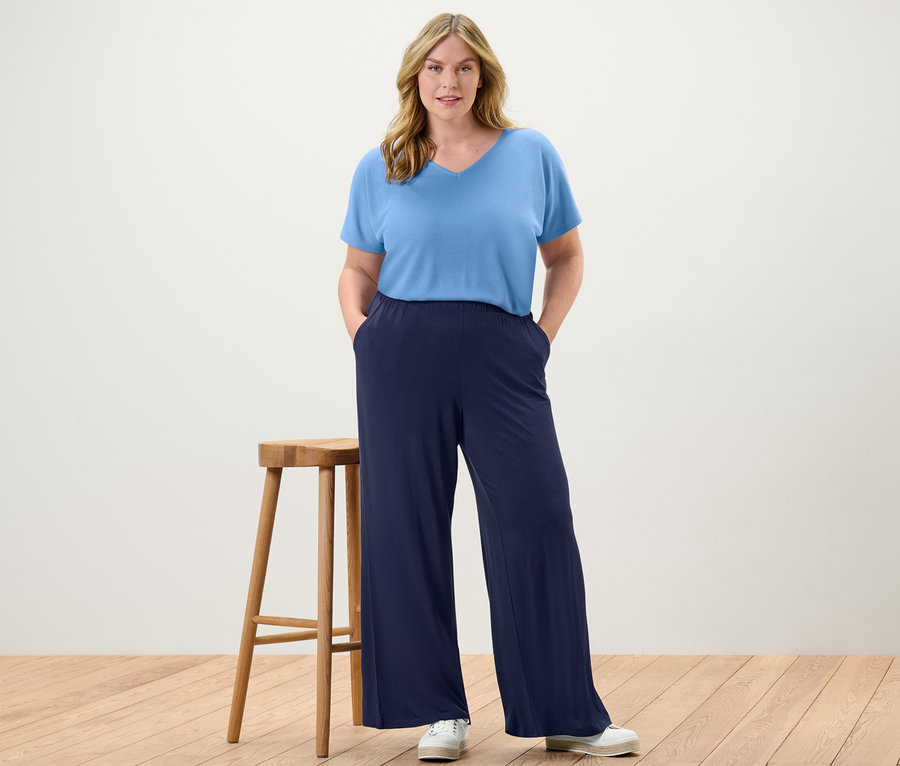 Žena stojí a má ruky vo vreckách tmavomodrých nohavíc v štýle palazzo a svetlomodrého štruktúrovaného trička.