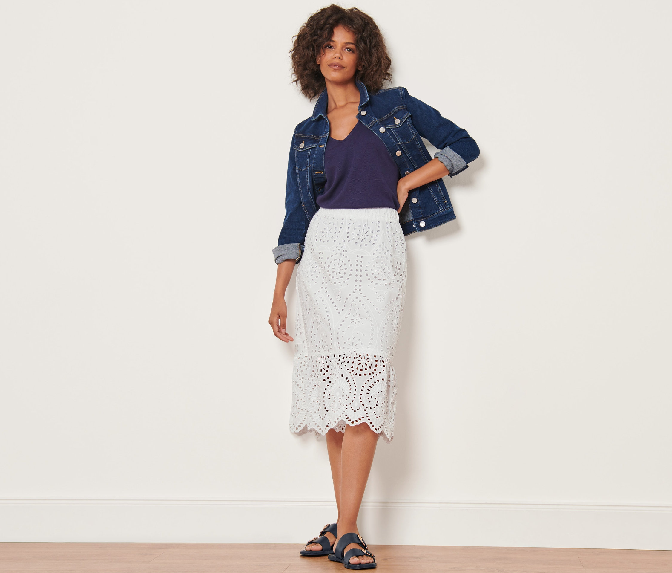 Žena pózuje v džínsovej bunde a bielej sukni s celoplošnou výšivkou.