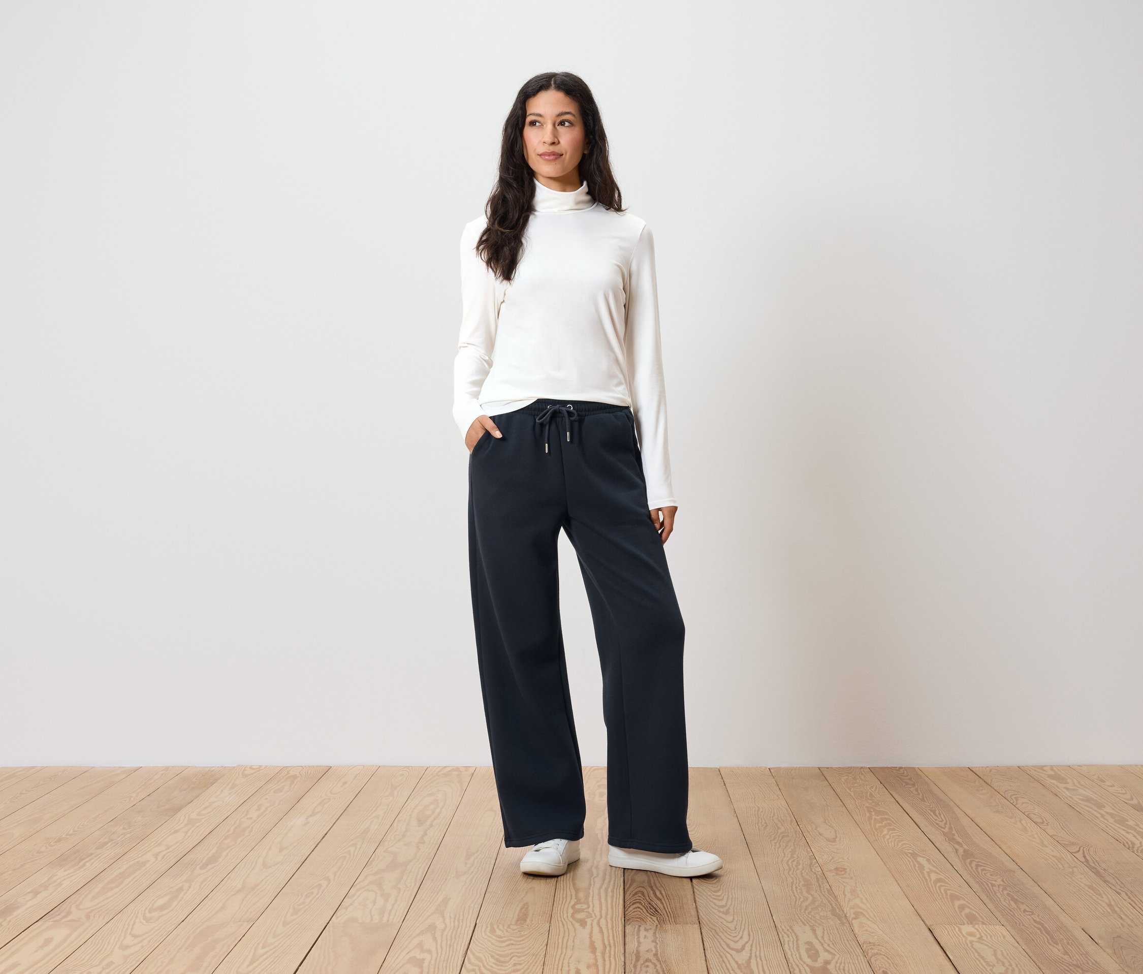 Žena stojí a má ruky vo vreckách tmavomodrých širokých nohavíc, biely rolák a biele tenisky.